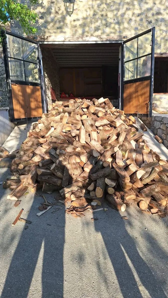 Pilha de lenha cortada em frente a uma garagem de pedra com portas abertas.