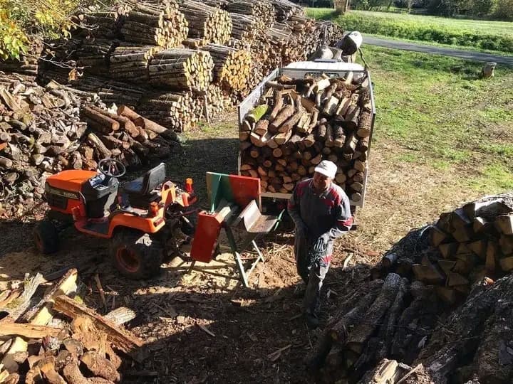 Homem operando rachador de lenha acoplado a trator laranja perto de pilhas de madeira.