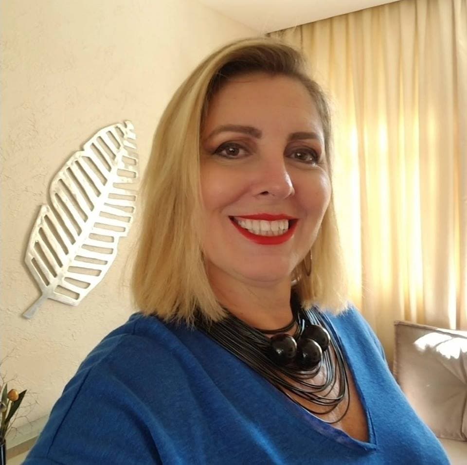 Mulher loira sorridente com blusa azul e colar preto em frente a uma parede decorada.