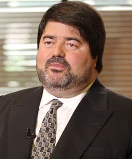 Homem de barba e cabelos escuros vestindo terno preto, camisa branca e gravata estampada.